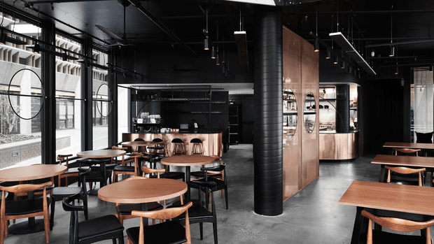 A dark, industrial-style cafe or restaurant interior with extensive black-painted ceilings, exposed services, suspended linear lighting, large factory-style windows, concrete flooring, and a prominent black cylindrical structural column. The space is furnished with numerous timber-topped dining tables and black chairs, while a copper-toned bar and service counter occupy the rear. Circular black-framed wall details and shelving add graphic features to the monochromatic fit-out.