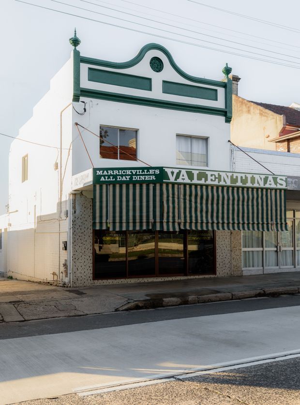 Street-level exterior photograph of a narrow two-storey heritage commercial building, currently a diner, with a white rendered facade, ornate curved parapet and green detailing, decorative finials, striped green-and-white awning, mosaic-tiled shopfront columns, and large ground-floor windows. The building fronts directly onto an urban sidewalk and street.