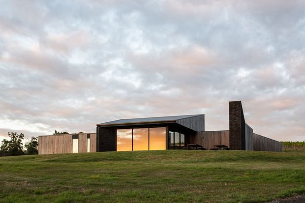 A low-slung contemporary house set in a broad grassy landscape, photographed at dusk. The building features dark vertically clad walls, extensive floor-to-ceiling glazing glowing warm orange from the interior, a projecting flat roof, and a tall dark masonry chimney or feature wall. Timber-clad wings extend on either side across the site.