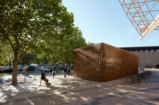 Exterior view of a contemporary urban architectural complex or public/commercial space. A rust-brown perforated metal cube-like pavilion sits on a paved plaza beneath mature trees, with outdoor café tables and chairs to the right. A larger masonry building and a distinctive exposed triangular steel canopy or roof structure are visible in the background, with pedestrians and parked vehicles along the street.