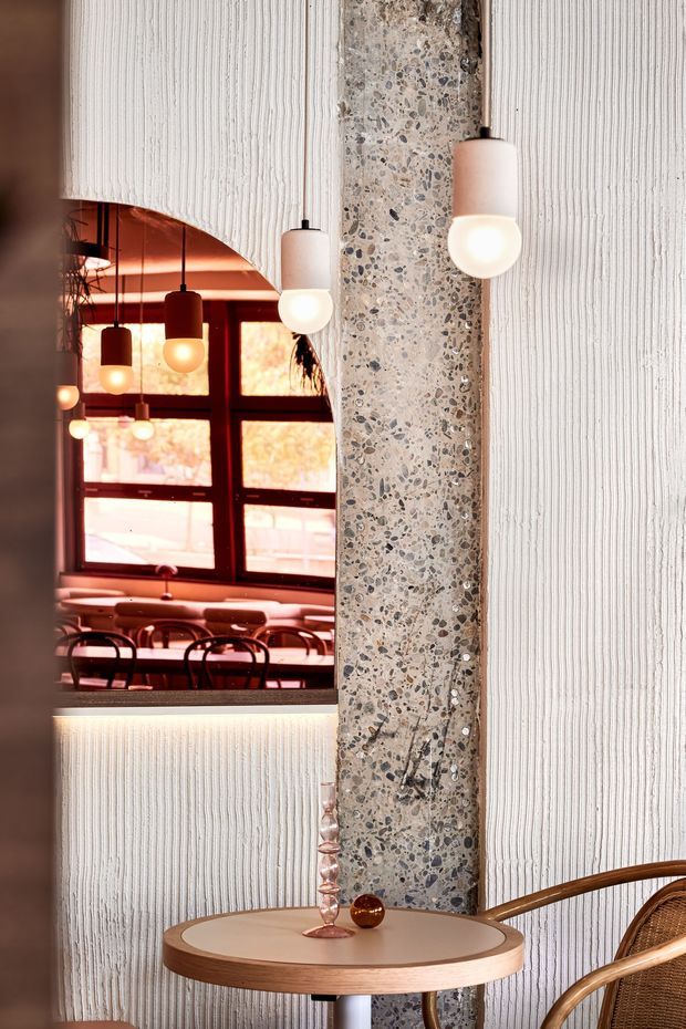 A warmly lit commercial interior featuring a textured white wall, a vertical terrazzo-clad structural column, and a curved opening framing a dining or café area beyond. Several hanging globe pendant lights glow above tables and bentwood-style seating. In the foreground, a small round wood-edged table holds decorative glassware, with a woven chair partially visible. The palette combines cream, warm terracotta, dark timber, and terrazzo tones.