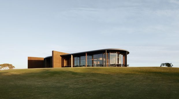 A low-profile contemporary building set on a gently sloping grassy site. The structure features a curved glazed frontage with extensive floor-to-ceiling windows, dark roof and framing elements, and warm timber-toned solid wall cladding. The composition is a minimalist architectural exterior photograph under a pale sky.