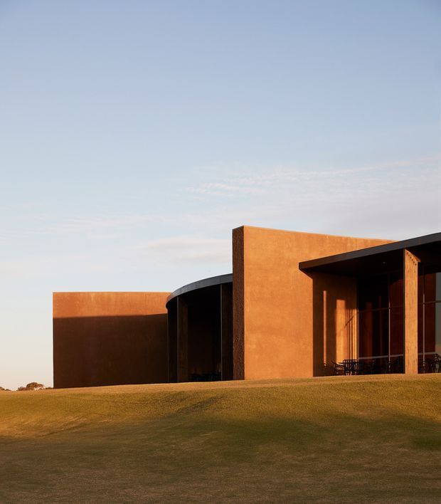 A low-slung contemporary building set into a gently sloping grass-covered site, photographed in warm late-afternoon light. The exterior features broad rust-toned rendered walls, a dark curved roof or canopy element, deep recessed openings, and a shaded covered terrace with timber-toned structural posts and outdoor seating. The composition emphasises the sculptural geometric facade against a pale blue sky.