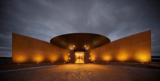 Dramatic twilight exterior of a contemporary entrance pavilion. Curved rust-coloured metal walls form a wide symmetrical forecourt, with a large oval, ribbed canopy and warm uplighting leading to a brightly glazed central doorway. The image has a moody, high-contrast architectural presentation.