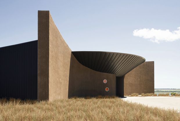 Exterior view of a sculptural contemporary building set in a sparse coastal or semi-arid landscape. The composition features tall textured earthen-toned walls, a dark vertically ribbed facade, and a dramatic curved cantilevered canopy with a ribbed underside forming a sheltered entry or outdoor space. Dry grasses, pale paving, and a distant body of water are visible under a clear sky.