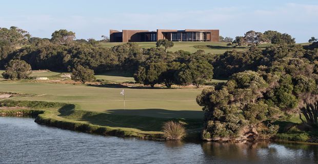Wide exterior landscape view of a low, horizontally composed contemporary building set on a gently elevated grassy site above a golf green and waterway. The building has dark brown and charcoal-toned rectangular volumes, extensive glazing, and a flat roofline, surrounded by mature native-looking vegetation. The golf course and water dominate the foreground.