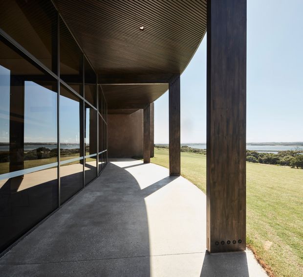 A covered exterior terrace or verandah along a highly glazed modern building, with slender dark timber-clad structural columns, a deep projecting roof with finely lined timber soffit, and a smooth concrete floor. Full-height glazing reflects the landscape and opens the building to broad lawns and a distant body of water. Strong sunlight creates dramatic shadows beneath the canopy.