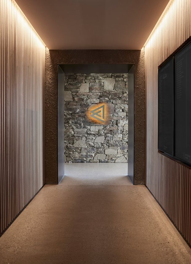 A narrow, warmly lit interior passageway or entry corridor with vertical timber slat wall lining on both sides, a textured earthen-toned ceiling and surrounding walls, and a polished concrete or terrazzo-like floor. At the end is a recessed opening framed in dark metal, revealing a rustic stacked-stone feature wall with an illuminated geometric emblem. A large dark display or screen panel is integrated into the right wall. The space has a refined rustic-industrial character with concealed linear lighting.