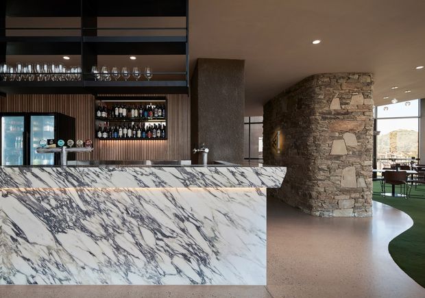 A sophisticated bar and hospitality-style interior dominated by a large veined stone counter. Behind the counter are dark timber slatted cabinetry, open shelving with glassware and bottles, under-counter refrigeration, and integrated lighting. A curved exposed-stone feature wall forms a sculptural partition beside a bright dining or lounge area with large glazing. The palette is warm and refined, combining dark joinery, pale stone, earthy flooring, and natural stonework.