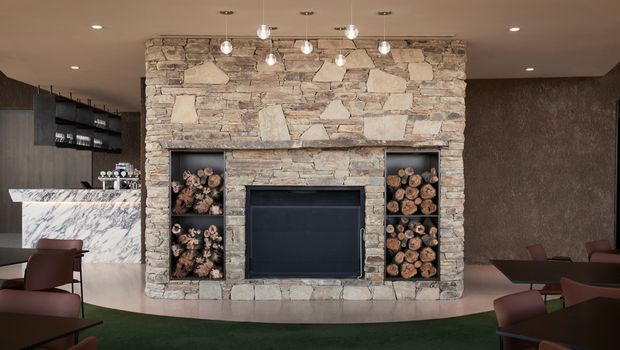 A sophisticated open-plan hospitality or lounge interior dominated by a large irregular natural-stone fireplace wall with a dark inset firebox and built-in log niches. A marble-clad bar or counter is visible to the left, with dark textured wall finishes, pendant globe lighting, and contemporary brown seating around the perimeter. The palette is warm and earthy with strong dark accents.