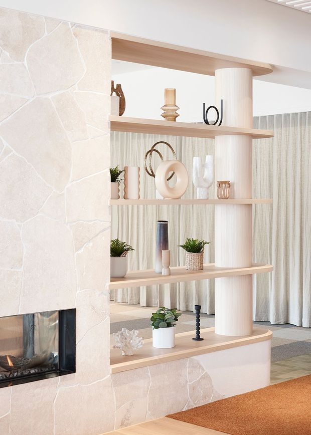 Bright, refined living room interior featuring a pale natural-stone fireplace surround with a built-in firebox, a curved timber open shelving partition, and a fluted cylindrical column. The shelves display neutral-toned ceramics, books, plants, and sculptural decor. Floor-to-ceiling sheer curtains, pale timber flooring, and a warm textured rug create a soft contemporary atmosphere.