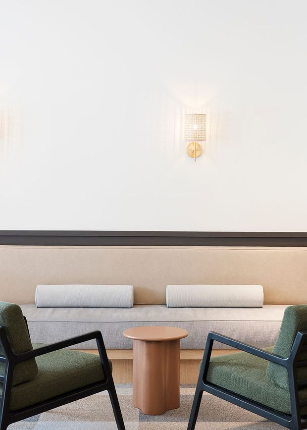 A refined, minimalist seating area with a long upholstered built-in bench against a white wall, two green upholstered armchairs with dark frames, and a small rounded timber side table. A warm wall sconce provides a subtle decorative accent, while layered neutral upholstery and carpeting create a calm, contemporary hospitality-like atmosphere.