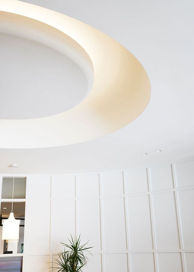 Bright contemporary interior featuring a large sculptural oval recessed ceiling feature with warm concealed lighting, a white coffered or panelled wall, recessed downlights, and a hanging pendant visible at the left. A potted plant adds a small natural element. The predominantly white space has a clean, refined, minimalist character.