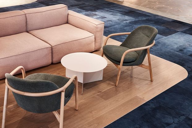 A contemporary lounge setting with a blush-pink sectional sofa, two dark upholstered timber-framed armchairs, and a round white coffee table arranged on a light timber platform. Large geometric dark blue-grey carpet tiles surround the seating area, creating a refined commercial or hospitality-style interior.