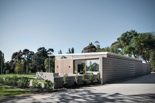 A low-profile modern single-storey building set within a landscaped suburban or park-like site. The structure features a flat roof, extensive glazing, pale rendered or concrete walls, and a prominent dark grey perforated block or screen wall extending along one side. A paved driveway or forecourt leads past the building, with planting and mature trees surrounding the site.