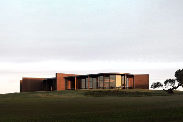 Low, single-storey contemporary building set prominently on a gently sloping grassy site. The composition features broad curved floor-to-ceiling glazing, dark slim window frames, warm reddish-brown solid wall volumes, and a shallow curved roofline. The surrounding landscape is open and rural, with a dramatic overcast sky and sparse sculptural trees.
