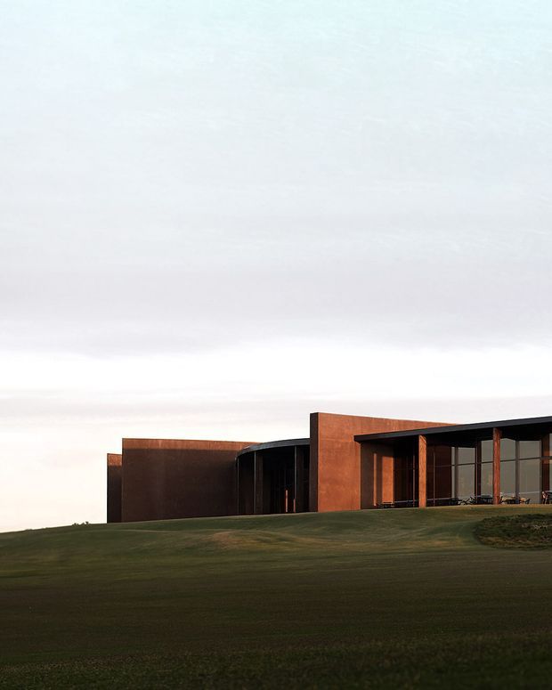 A low, contemporary building set into a broad grassy landscape beneath an overcast sky. The facade combines deep rust-brown panels with dark framing, expansive glazed openings, and a curved recessed entrance or wing. The composition emphasizes horizontal massing, strong geometric volumes, and a restrained connection to the surrounding lawn.