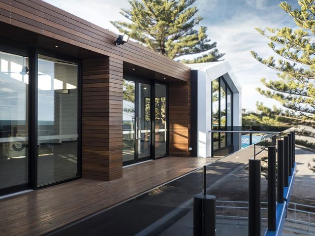 Modern coastal building exterior with dark timber cladding, extensive black-framed glazing, and a white angular glazed extension. The view is from an elevated deck or walkway with black balustrades, overlooking a bright blue pool and mature coastal trees. The architecture has strong horizontal lines, clean geometric forms, and a contemporary outdoor connection.