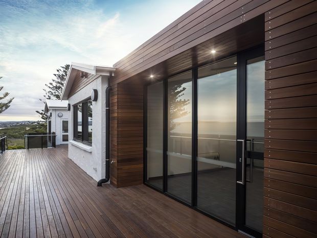 A modern coastal-facing home exterior viewed from a broad timber deck. The composition features dark horizontal timber cladding, black-framed floor-to-ceiling glazed sliding doors, a recessed timber-lined soffit with downlights, and an adjoining white rendered gabled section. The deck overlooks a distant body of water and rolling landscape.