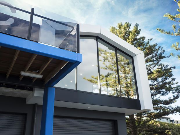 Exterior view of a contemporary multi-level building, showing a bold blue cantilevered balcony or awning, dark-framed floor-to-ceiling glazing, a white angular projecting upper volume, and a garage door below. The facade is set against mature trees and a bright sky.