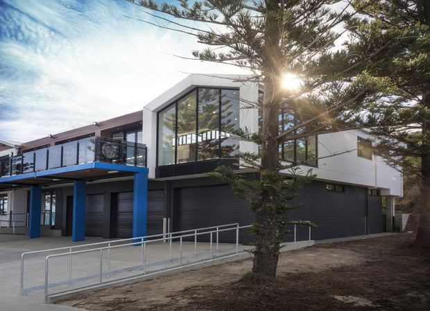 Exterior view of a contemporary multi-level building with a bold white and dark-grey facade, large floor-to-ceiling glazed gable-like section, blue structural columns and balcony trim, multiple dark garage doors, and an accessible ramp with metal handrails. Large established trees partially screen the building.
