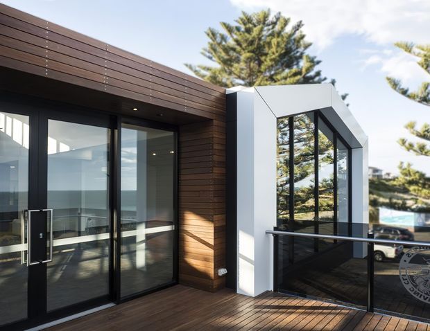 Contemporary building exterior with a timber-clad entrance and deck, black-framed glazed doors, and a projecting white architectural volume containing tall windows. The image is taken from an outdoor terrace or balcony, with strong reflections and glimpses of the surrounding landscape and water beyond.