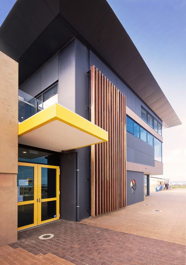 Exterior architectural photograph of a contemporary multi-storey building entrance. The facade combines dark grey panel cladding, extensive blue-tinted glazing, a projecting black roof canopy, warm vertical timber screening, and a vivid yellow entrance canopy and door surround. A brick-paved forecourt and open paved walkway are visible, with a bright coastal-looking horizon in the distance.