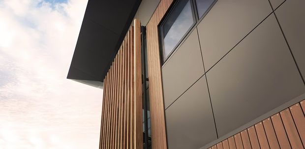 Upward-looking close-up of a contemporary building facade featuring vertical timber-look screening, large dark-framed windows, smooth light-grey panel cladding, and a broad projecting roof or canopy against the sky.