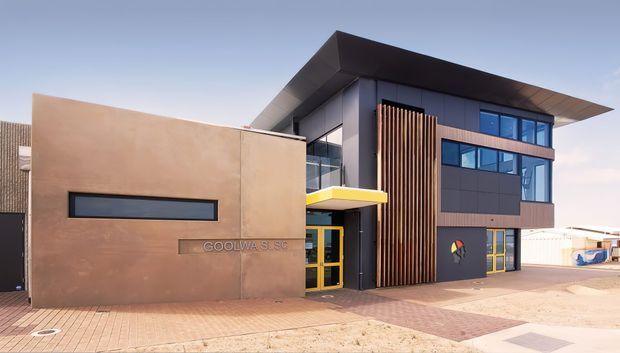 Contemporary two-storey commercial building with a strong geometric facade, dark cladding, warm timber vertical screening, extensive blue-tinted glazing, and a projecting flat roof. A smooth tan rendered entry volume features narrow horizontal glazing and signage, while a bright yellow canopy and matching door frames provide accents. The building fronts a paved pedestrian area under a clear sky.