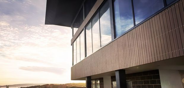 Contemporary multi-level building exterior photographed at sunset, featuring extensive floor-to-ceiling glazing, dark vertical cladding, a projecting upper volume, and a lower level supported by dark structural columns. The building sits in an open landscape with distant low hills and sky visible beyond.