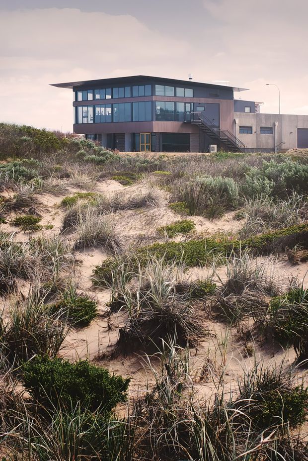 Exterior view of a contemporary multi-level building set behind sandy coastal dunes. The building features broad floor-to-ceiling glazing, dark metal window frames, a low overhanging flat roof, dark cladding, an external stair, and an attached concrete service volume.