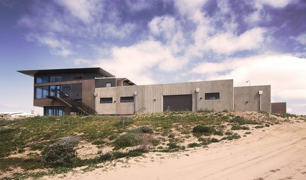 Exterior view of a contemporary, low-profile beachside or dune-site building with a rectilinear form. The structure combines a darker two-storey glazed wing with an external stair and a long, single-storey ribbed-cladding volume containing a large garage-style door and narrow horizontal windows. It is set among sandy dunes with sparse coastal vegetation under a dramatic cloudy sky.