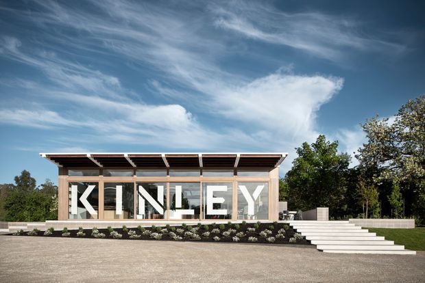 Single-storey contemporary pavilion-like building with extensive floor-to-ceiling glazing, a broad flat projecting roof with exposed dark rafters, and large white "KINLEY" lettering across the glazed facade. The structure sits behind a landscaped garden bed with broad steps leading to a raised deck or entry platform. Mature trees and an open paved forecourt surround it under a bright blue sky.