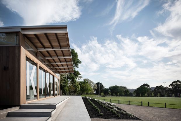 A modern low-rise building viewed from outside, featuring extensive floor-to-ceiling glazing, dark timber cladding, a broad projecting roof canopy with exposed timber soffit, and concrete terraces and steps. The building opens onto a landscaped garden and large open lawn, with a rural or suburban outlook.
