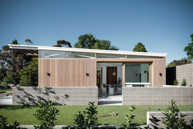 A low-slung contemporary single-storey house viewed from the landscaped front yard. The facade combines vertical timber cladding, pale grey concrete blockwork, expansive glazed openings, and a shallow mono-pitched roof with high clerestory-style glazing. A recessed entry and covered outdoor area open onto a neat lawn and garden.