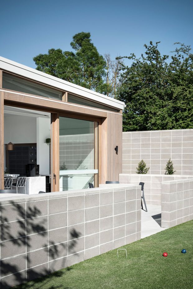 Exterior view of a compact modern pavilion or extension opening onto a private courtyard. The building has a low white roofline, extensive glazing and warm timber-framed sliding doors, with a pale grey concrete-block boundary wall enclosing a paved outdoor area. A small lawn and garden planting are visible in the foreground, and a kitchen or living area can be seen through the open glazing.