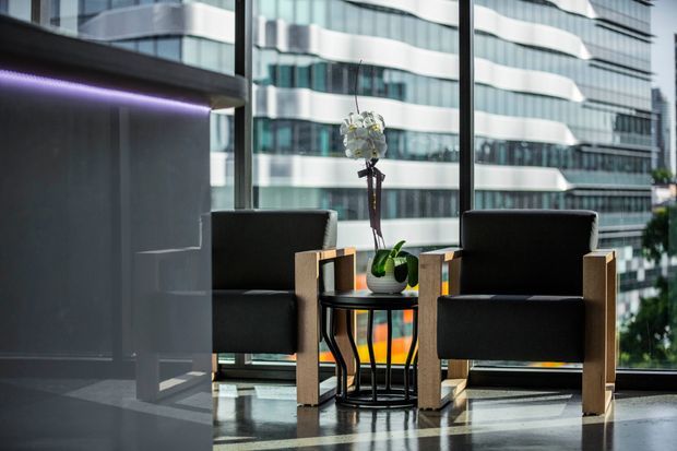A contemporary high-rise interior lounge or waiting area with two dark upholstered armchairs positioned beside a small side table and potted orchid. Full-height glazing overlooks a dense urban setting with modern office buildings. The space features a polished reflective floor, dark wall surfaces, timber-toned chair frames, and subtle purple linear lighting.