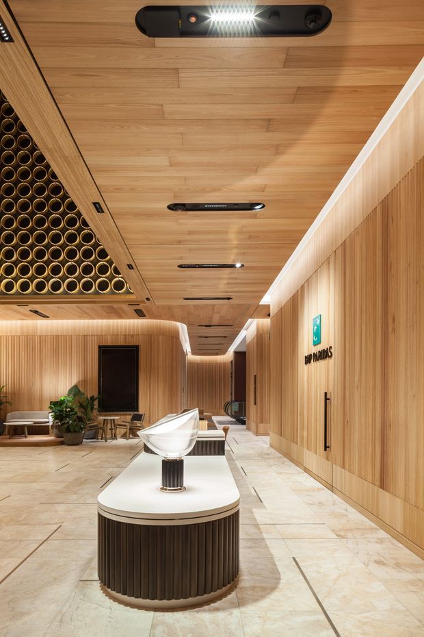 A refined commercial interior lobby or reception corridor featuring extensive vertical timber wall panelling and a matching timber-lined ceiling with recessed linear lighting. A sculptural white reception or display island with a dark fluted base occupies the foreground, while a lounge seating area and plants sit in the background. The space has polished pale stone-look flooring, rounded corners, a decorative circular timber ceiling feature, and clear wayfinding signage. The overall design is warm, contemporary, and high-end.