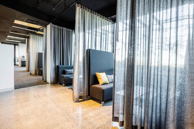 A contemporary commercial interior featuring a series of semi-private lounge or meeting booths screened by full-height sheer curtains. The space has a dark exposed ceiling with suspended linear lighting and services, polished speckled concrete flooring, carpeted areas beyond, and deep upholstered seating in charcoal and blue tones with occasional yellow cushions. Daylight filters through large windows behind the curtains.