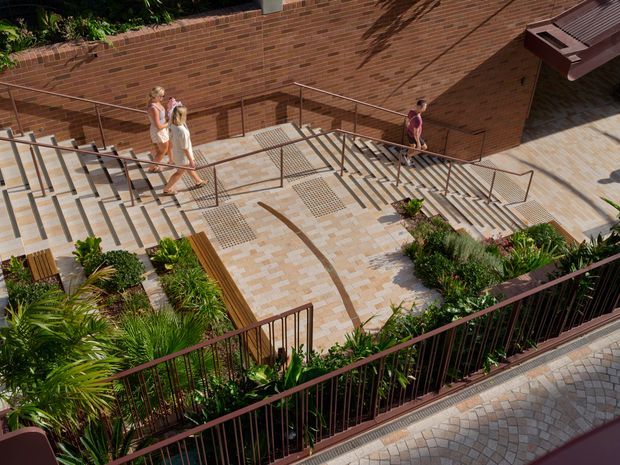 A landscaped, accessible outdoor stairway and ramp precinct beside a long red-brick building wall. Broad pale patterned paving, multiple stainless-steel handrails, planted garden beds, timber bench seating, and dark metal balustrades create a well-designed public or commercial circulation space. Several pedestrians use the stairs and ramp.