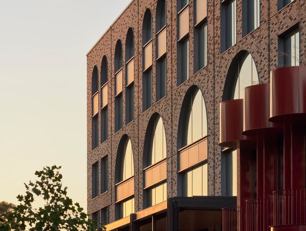 Close-up exterior architectural view of a multi-storey urban building with a warm reddish-brown brick facade, repeated tall arched window openings, dark window frames, projecting rectangular surrounds, and sculptural deep burgundy curved balcony or facade elements. The building is photographed in warm late-day light, with a small amount of tree canopy at the lower edge.