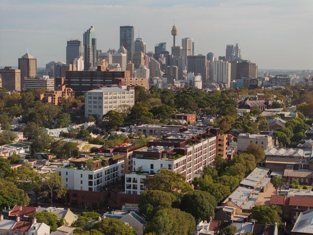 A wide elevated cityscape view showing a dense modern urban skyline in the background, including a prominent communications tower, with leafy suburban neighbourhoods, low-rise commercial buildings, and apartment blocks in the foreground. The scene is photographed in warm daylight with a hazy atmosphere.
