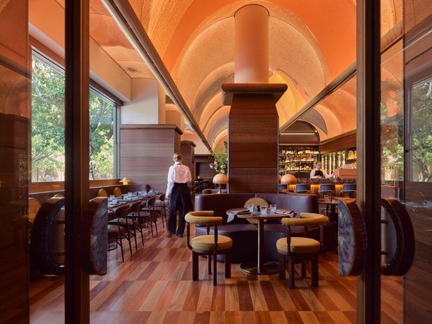 A richly detailed restaurant interior viewed through glazed entry doors. The space features a long central axis, extensive warm timber wall panelling and flooring, dark framed glass partitions, banquette seating, round dining tables, and a dramatic vaulted barrel ceiling with integrated warm orange lighting. A prominent central timber-clad column or fireplace structure anchors the dining room, with staff and additional seating visible deeper in the space.