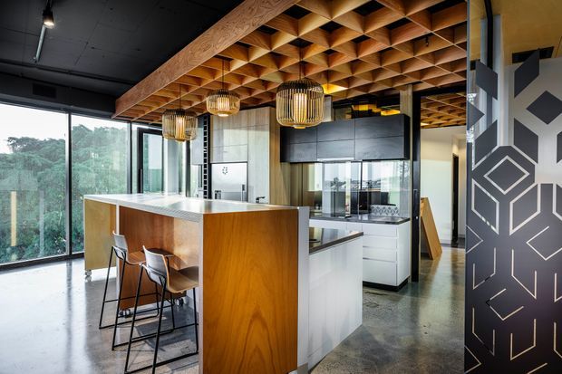 Contemporary kitchen with a large timber island and integrated seating, white cabinetry, dark overhead joinery, polished concrete flooring, full-height glazing, and a distinctive geometric timber ceiling with pendant lights. A patterned black feature wall and reflective surfaces add visual contrast.
