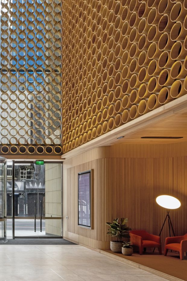 A sophisticated commercial lobby or hotel reception foyer with a tall atrium, warm timber wall panelling, pale stone flooring, and a striking suspended or screened feature made from repeating circular timber elements. The space includes a glazed entrance, framed wall display, and a lounge nook with rust-red upholstered lounge chairs, potted plants, and a sculptural floor lamp. The design combines warm natural materials with contemporary architectural detailing and abundant daylight.