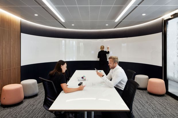 A contemporary commercial meeting and collaboration room with a long white conference table, ergonomic black chairs, and several rounded upholstered stools. The room features a curved white writable wall with a dark horizontal band, warm vertical timber wall panelling, integrated linear and recessed lighting, a suspended grid ceiling, and dark patterned carpet. A person is writing on the wall, reinforcing the room’s function as a collaborative workspace.