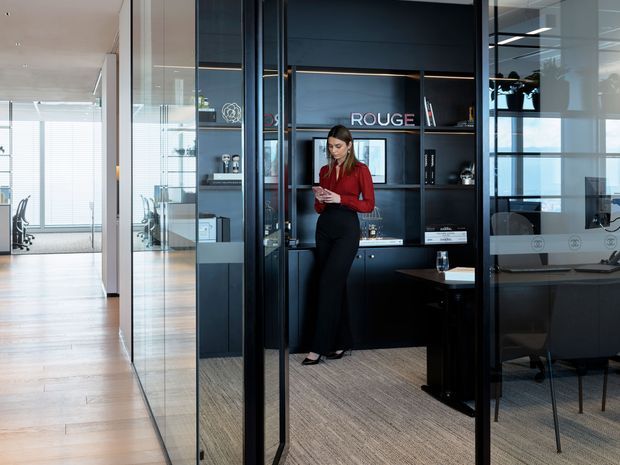 A contemporary office interior viewed through a glazed meeting-room partition. The space features dark built-in cabinetry and shelving, black-framed glass walls and doors, a carpeted meeting room, timber flooring in the corridor, and bright open-plan work areas beyond. A woman stands near the cabinetry, with desks and office chairs visible through the glass.