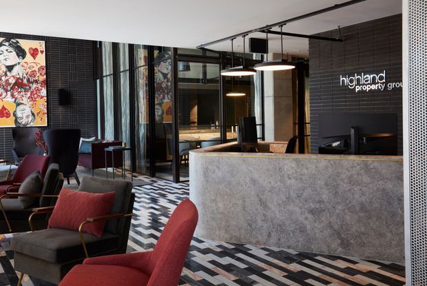 A sophisticated commercial reception area with a curved, textured stone-look reception desk, black tiled feature wall bearing the Highland Property Group logo, black-framed glazed partitions, pendant lighting, patterned geometric flooring, and contemporary lounge seating. Artwork and dark timber or black finishes add a refined, dramatic character.