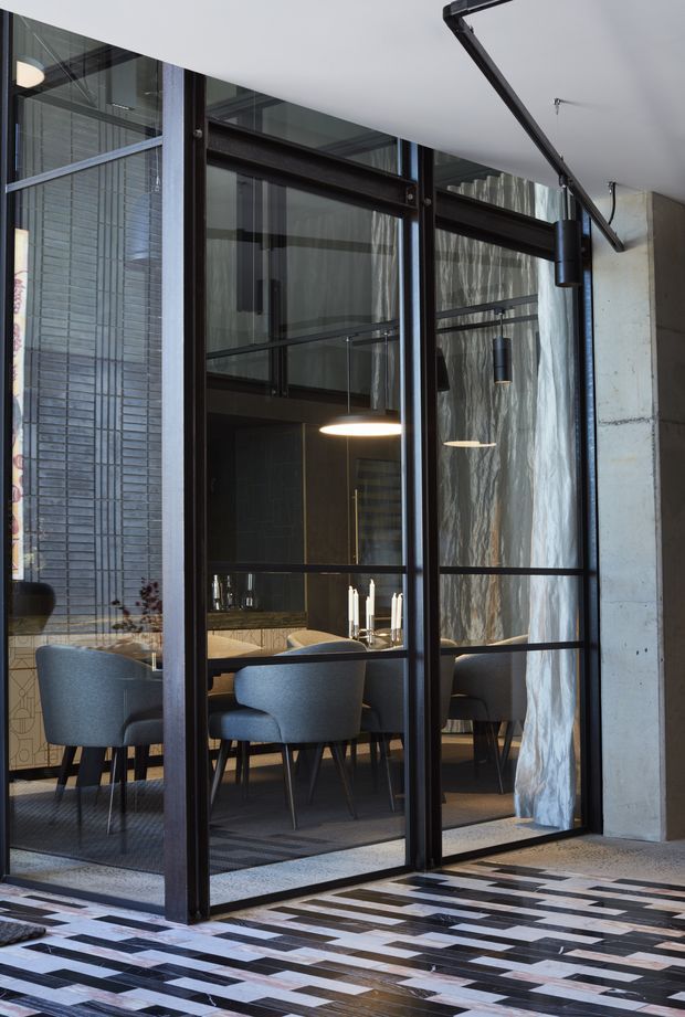 A dramatic interior view through black steel-framed glazed doors into a refined dining area. The room features a timber dining table, upholstered dining chairs, pendant lighting, dark cabinetry or wall finishes, and floor-to-ceiling curtains. A patterned timber floor continues in the foreground, while a raw concrete wall and exposed industrial-style door hardware frame the space.