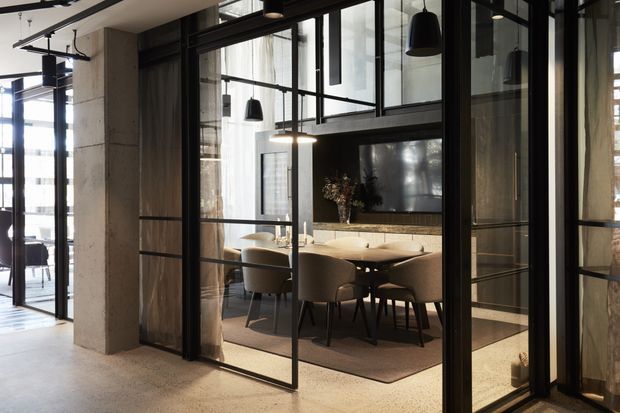 A refined contemporary dining room viewed through black-framed glazed partitions. The space features a long dining table with upholstered chairs, pendant lighting, a wall-mounted television, dark timber or stone detailing, and concrete-toned structural columns and flooring. Floor-to-ceiling glazing and internal steel-framed doors create a strong connection with the adjacent open-plan area.